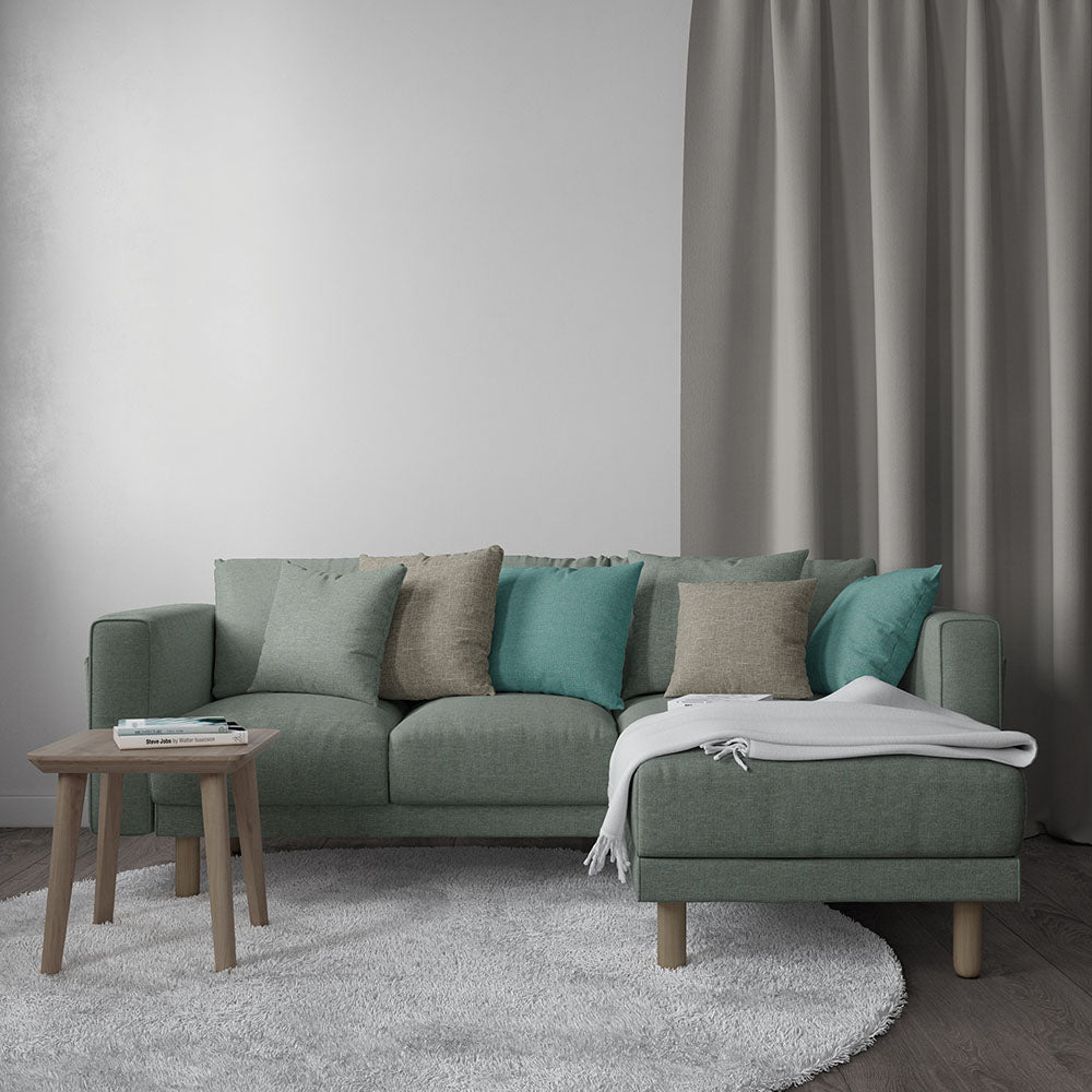 Green sage sectional sofa with chaise, teal/beige cushions, white throw, wooden legs, on a white rug under grey curtains.