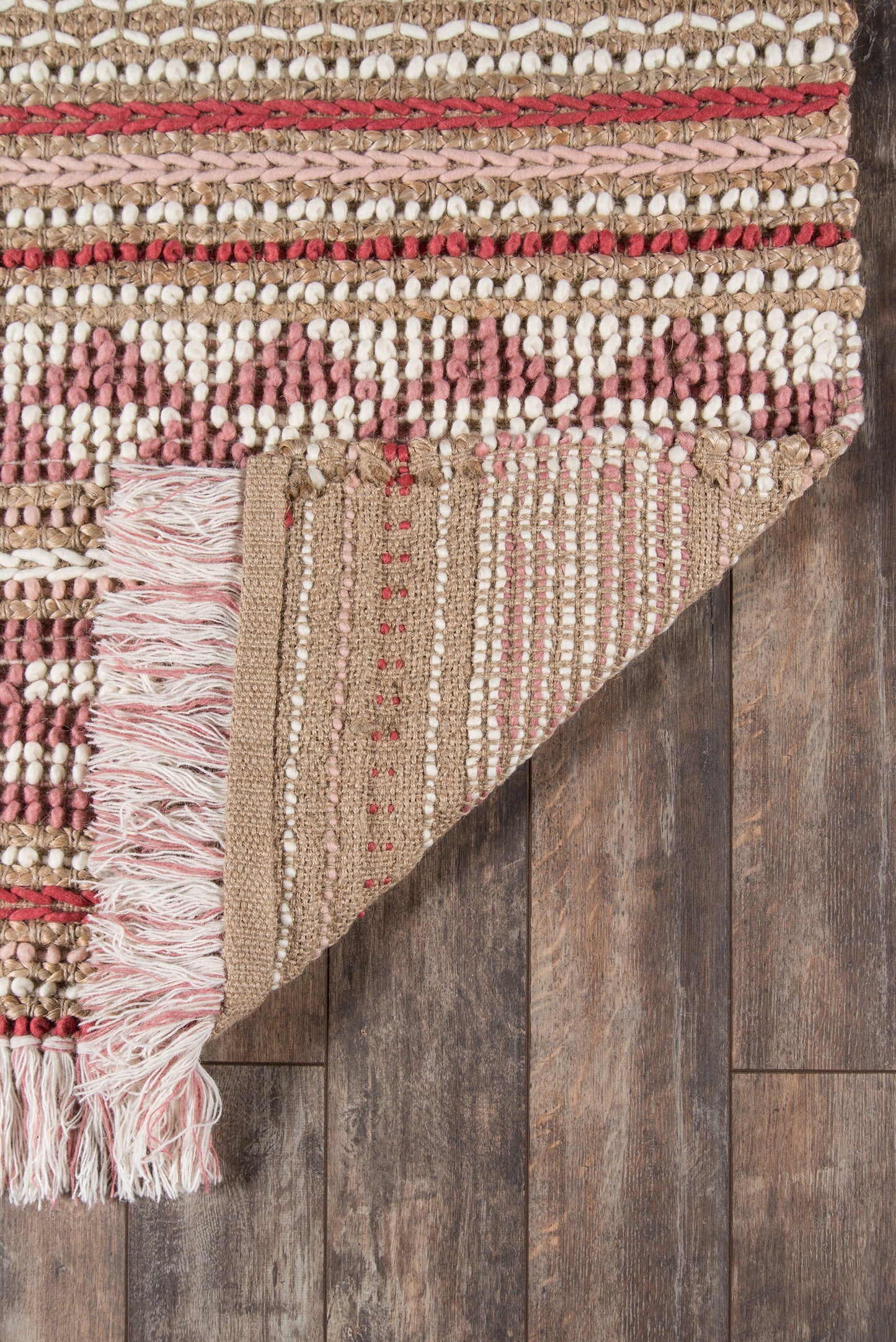 Handwoven beige rug with cream and pink stripes, textured weave, fringe tassels along edge, laid on dark wooden floor.