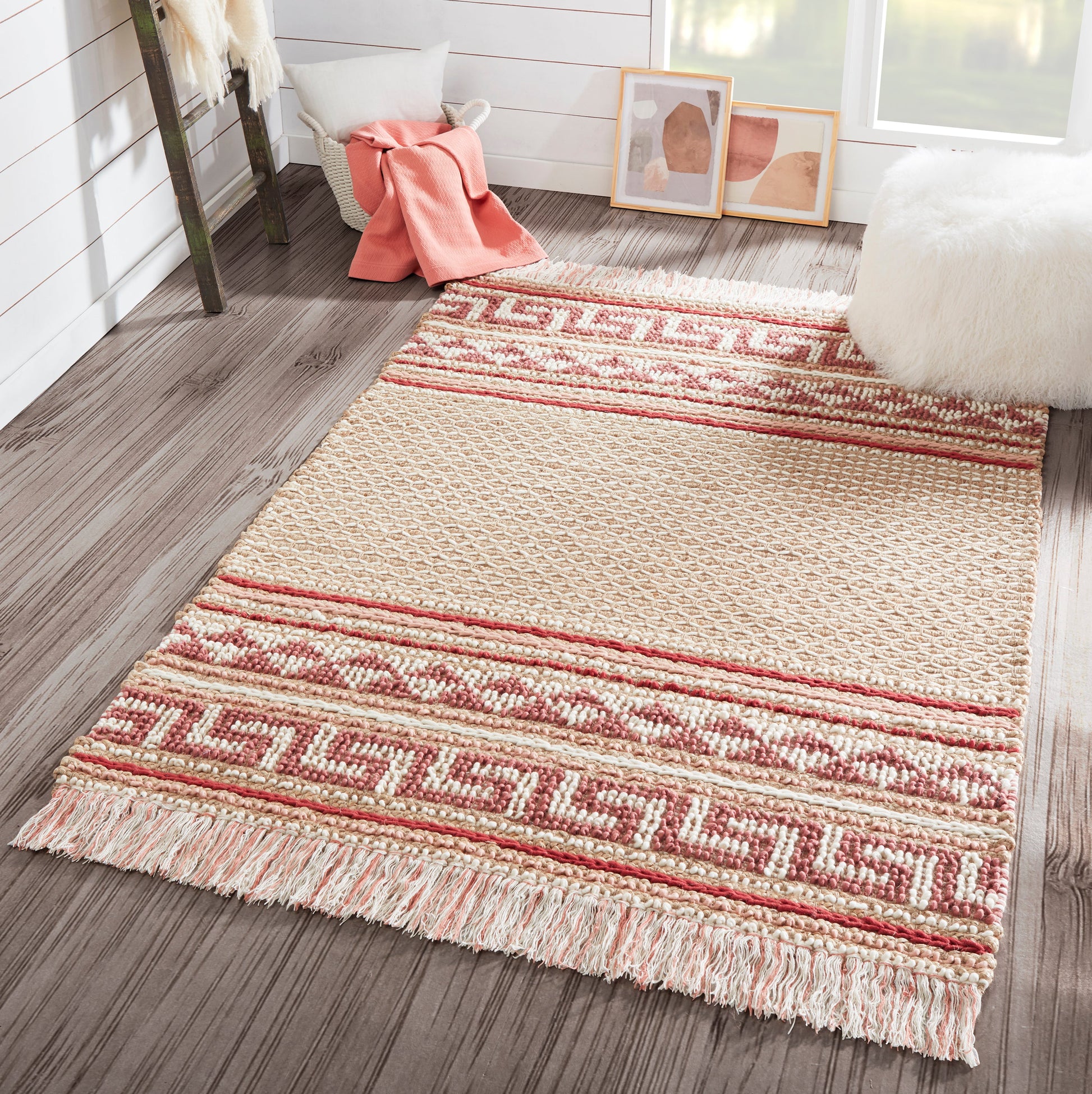 Beige & red geometric woven area rug with fringe ends on wood floor in a cozy corner with pink throw and fluffy ottoman.