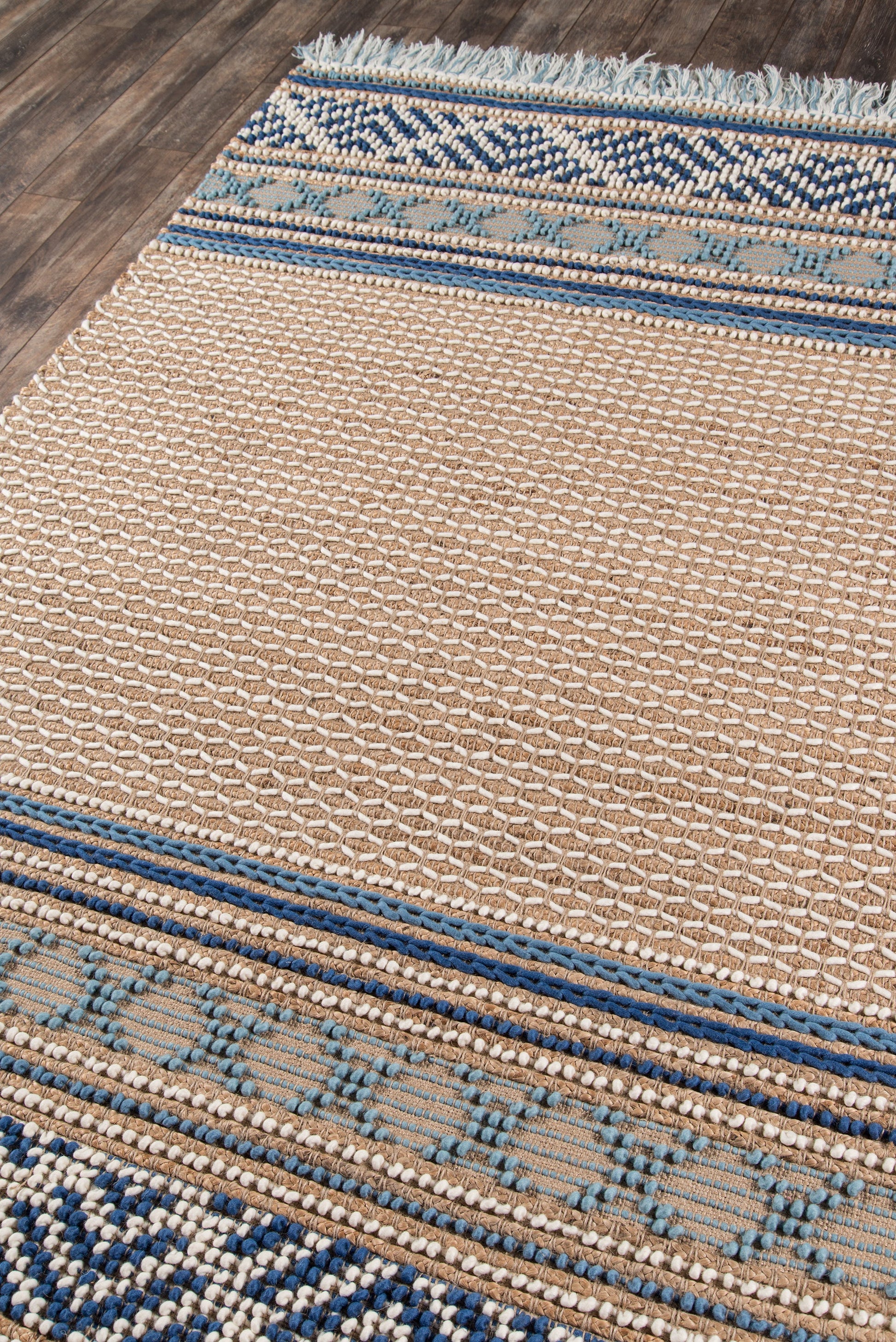 Rectangular beige flatwoven rug with blue geometric borders and fringe on a wooden floor.