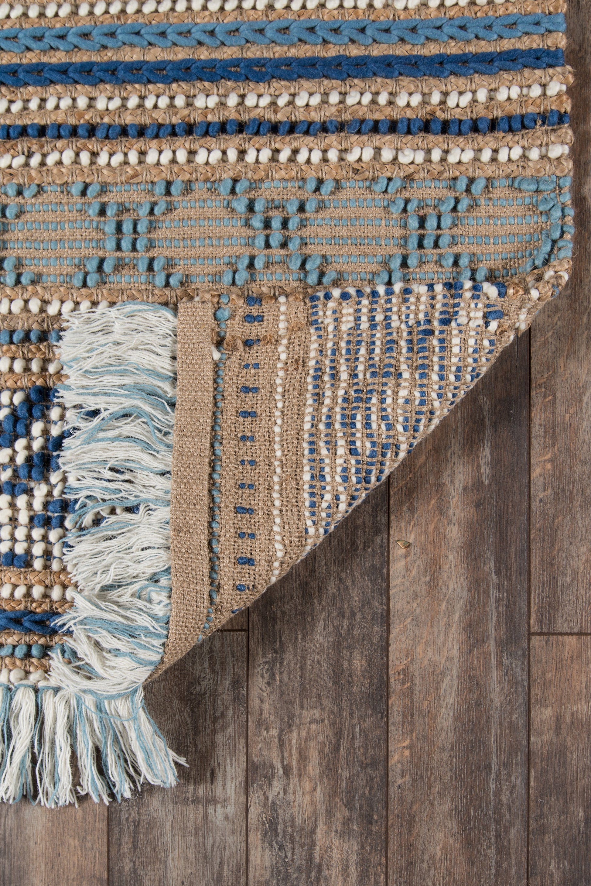 Boho woven rug in blue and beige stripes with geometric pattern, fringe tassels, folded corner showing underside on dark wood floor.