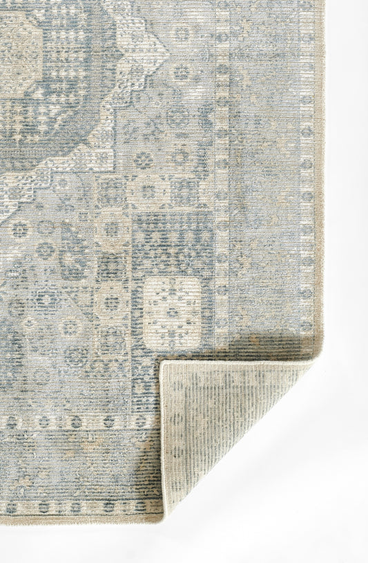 Blue-grey distressed oriental rug with intricate geometric floral pattern; folded corner reveals beige underside.