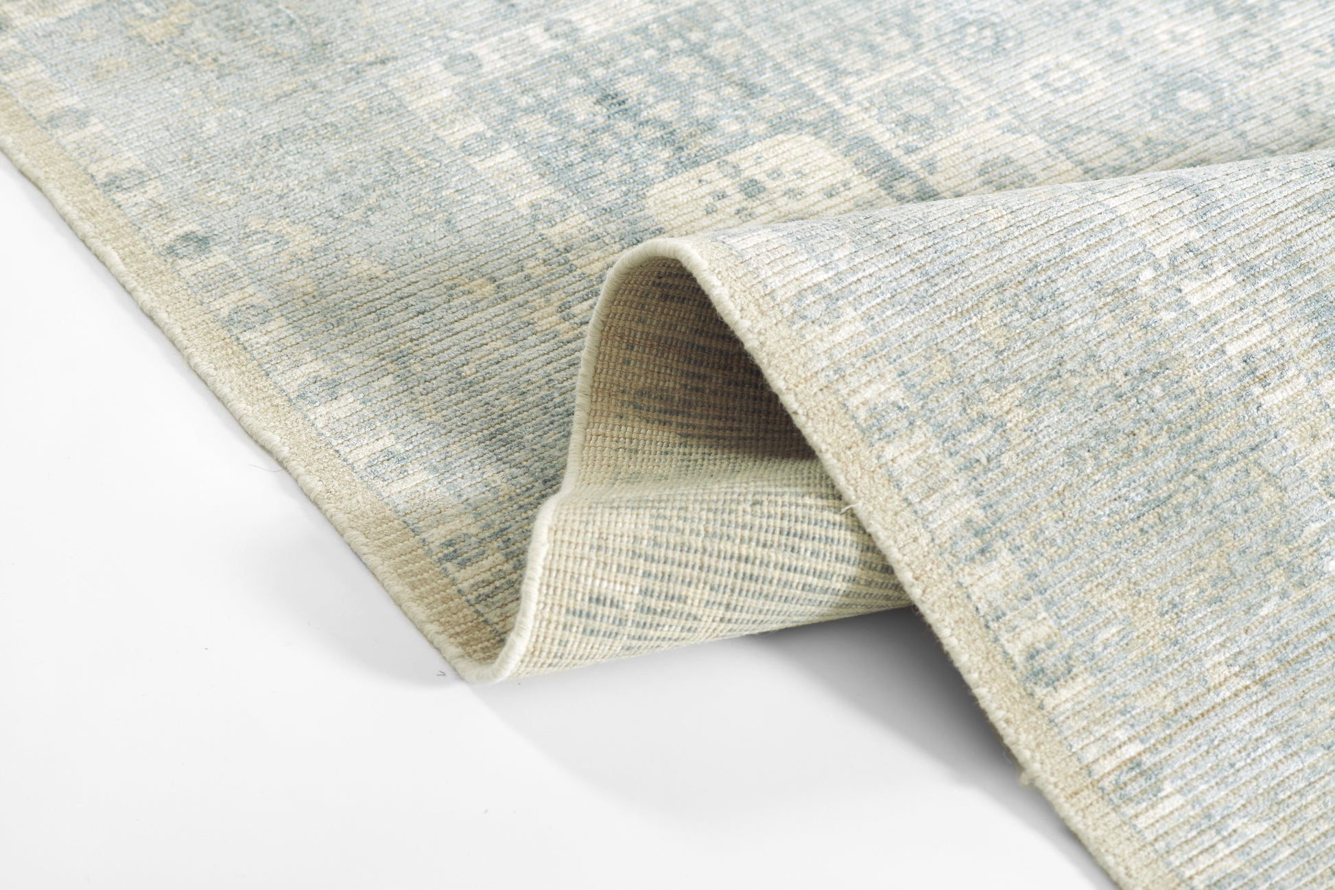 Blue-gray patterned rug with distressed motifs and soft woven texture; folded corner reveals a light underside.