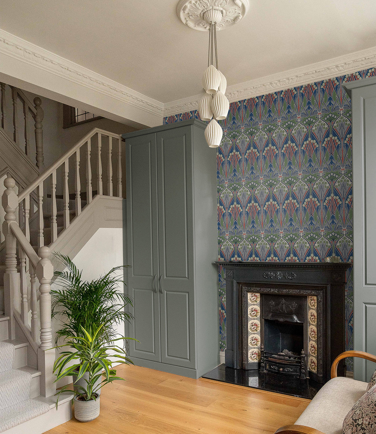 Traditional living room with blue-gray built-ins, ornate black tiled fireplace, patterned wallpaper, staircase, plant.
