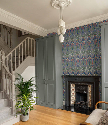 Traditional living room with blue-gray built-ins, ornate black tiled fireplace, patterned wallpaper, staircase, plant.