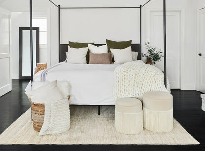 Natural woven rug (5' x 8') in a bright minimalist bedroom scene, featuring a black metal four-poster bed, white linens, green and beige pillows, a cream knit throw, wicker ottomans, and a woven basket.