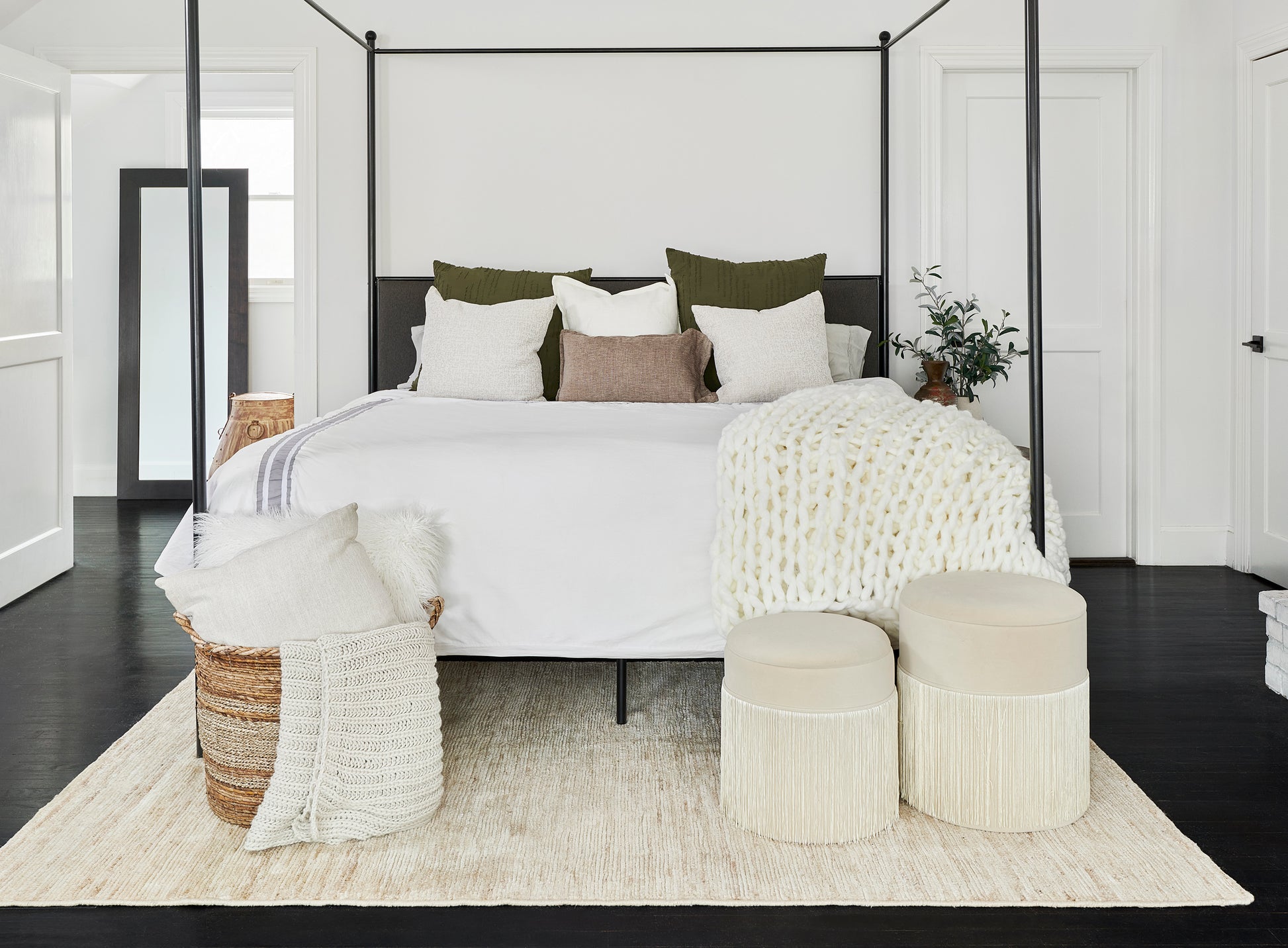 Canopy bed with black metal frame, white bedding, olive & cream pillows, cream knit throw, woven poufs, plant, beige rug.