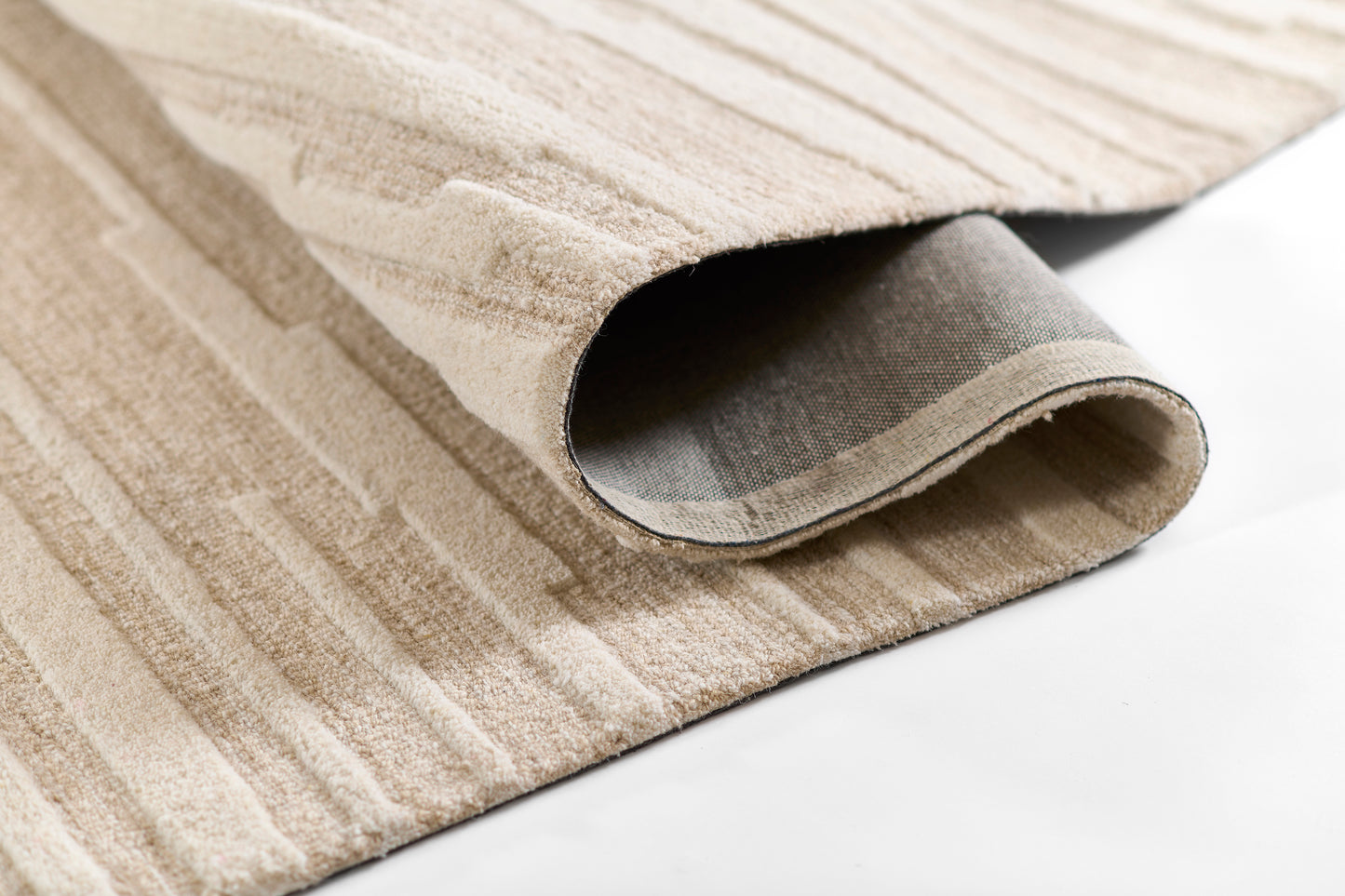 Beige striped area rug with soft pile, partially rolled to show gray backing and geometric line pattern.