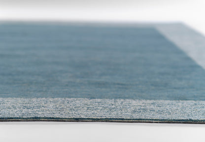 Close-up of a rectangular blue area rug with a light gray border and textured surface.