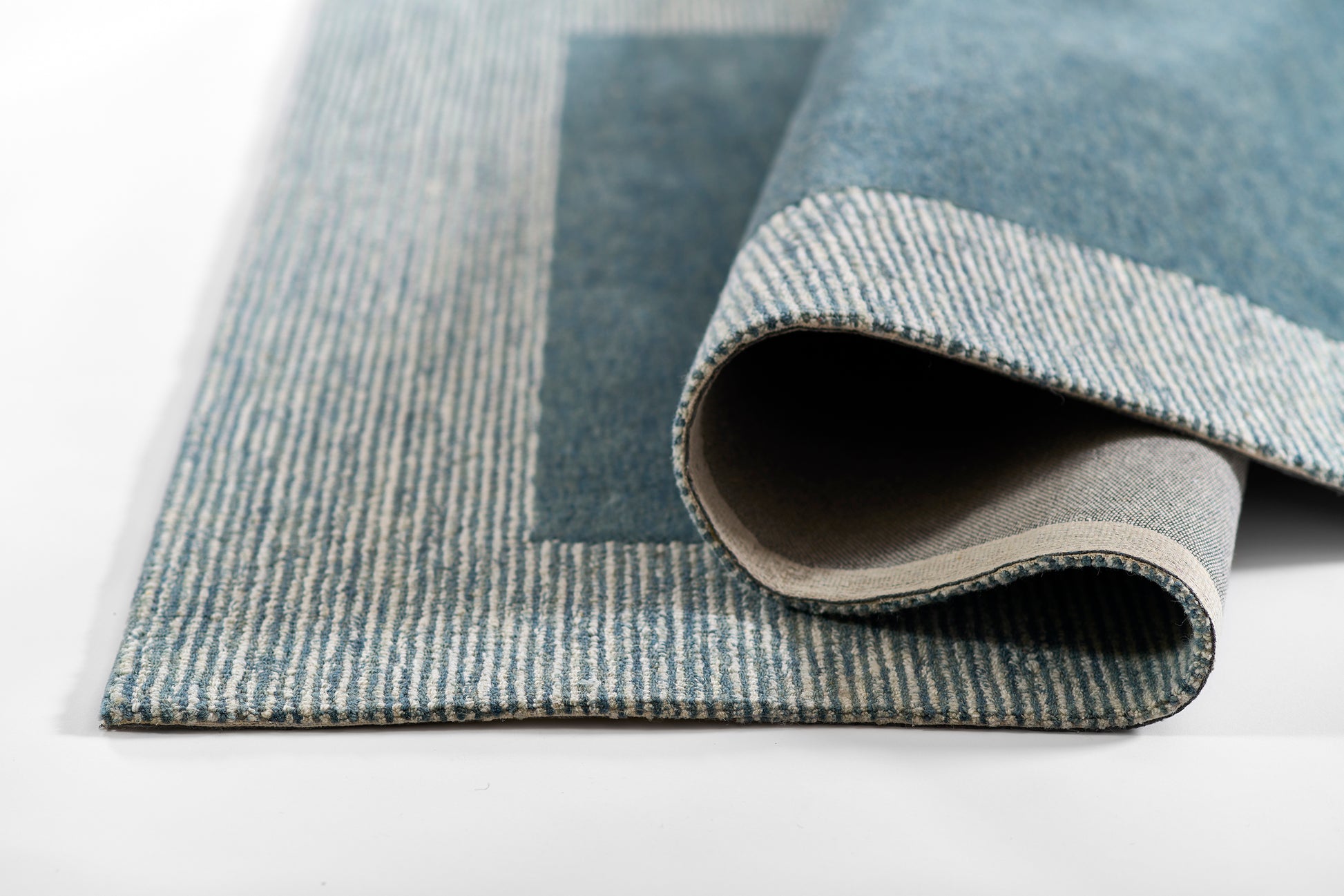 Blue denim-style rug with light striped border, rolled to show beige backing; modern woven floor covering.