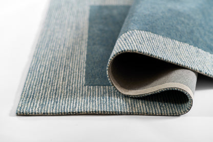 Blue denim-style rug with light striped border, rolled to show beige backing; modern woven floor covering.