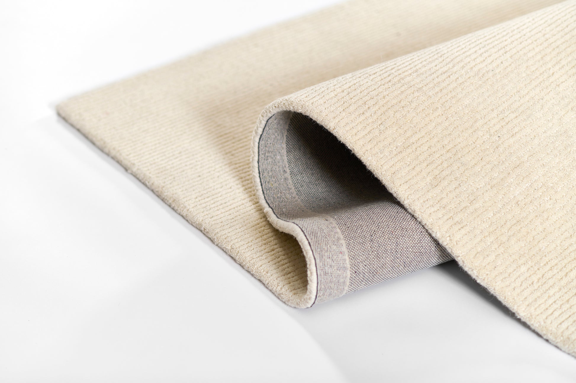 Beige woven rug with a soft ribbed texture and rolled edge, showing ivory top and gray underside.