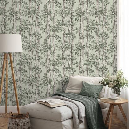 Beige chaise with green cushions and throws in a room featuring green trees botanical wallpaper, a wooden tripod floor lamp, and a round wood side table with a vase and a book.