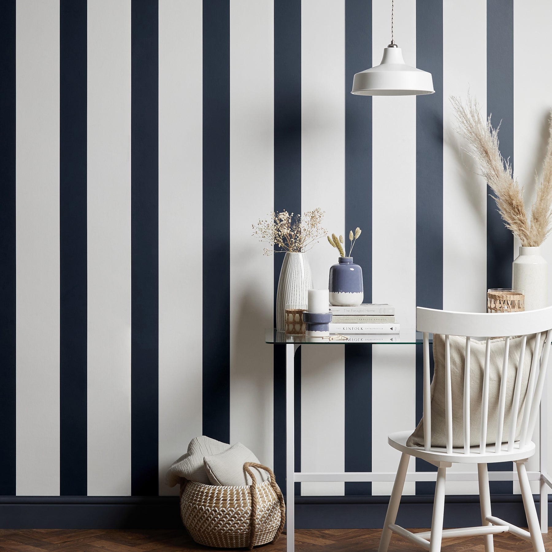 Blue and white vertical striped wallpaper with a glass table, white chair, ceramic vases, books, and a woven basket.