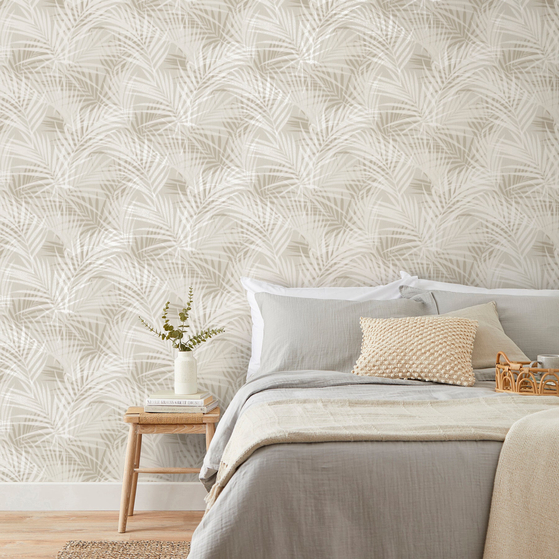 Neutral palm-pattern wallpaper in beige with gray bedding, beige textured cushion, wooden nightstand, vase and books.