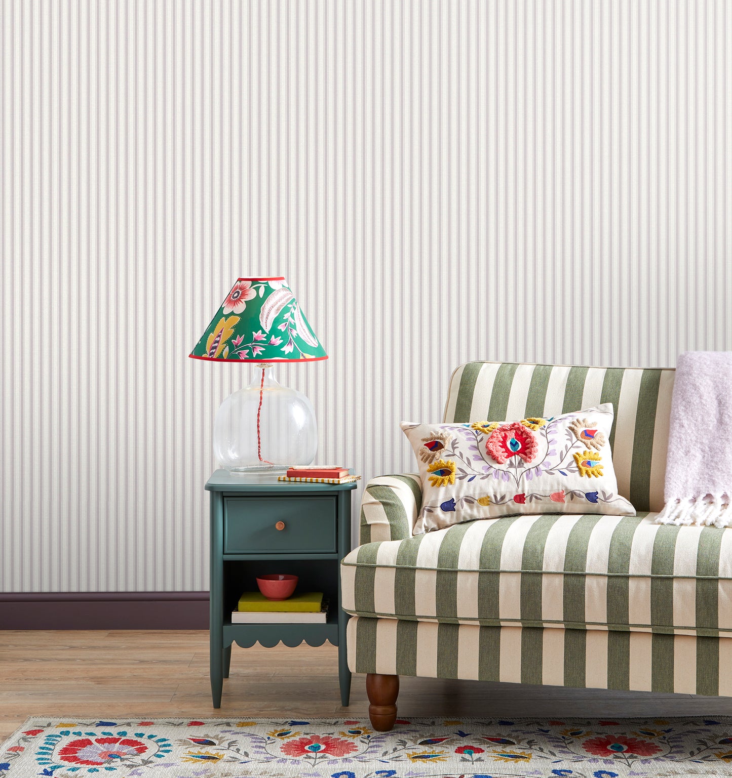 Teal painted end table with a clear glass base lamp & a bright floral shade beside a green-and-cream striped sofa; floral cushion & patterned rug.