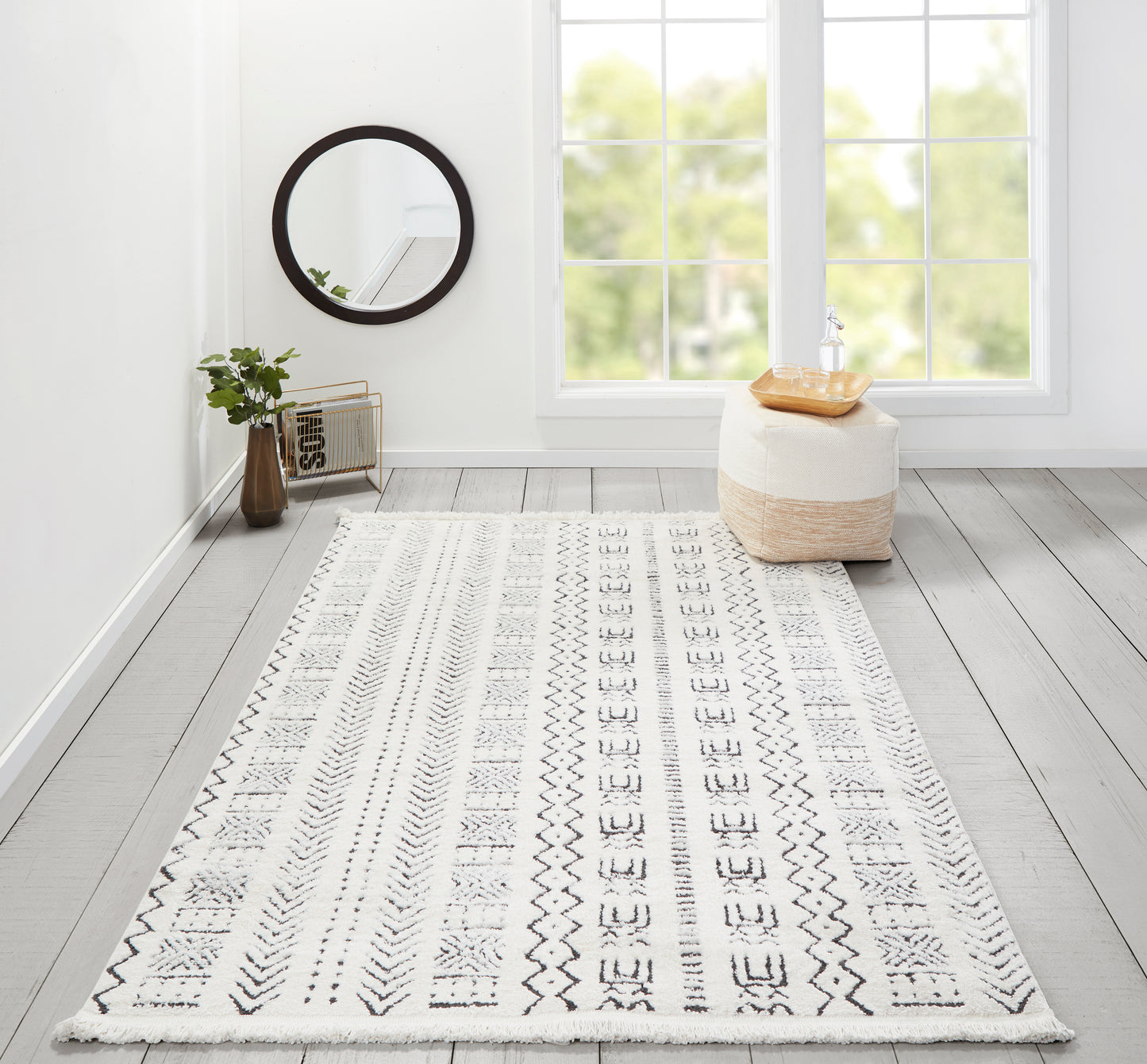 White geometric runner rug with fringe (2' x 8') on a light gray wood floor, round black-framed mirror, plant & beige ottoman with tray.