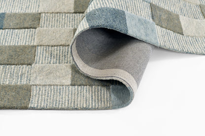 Blue-gray patchwork area rug with woven square blocks; rolled edge reveals lighter underside.