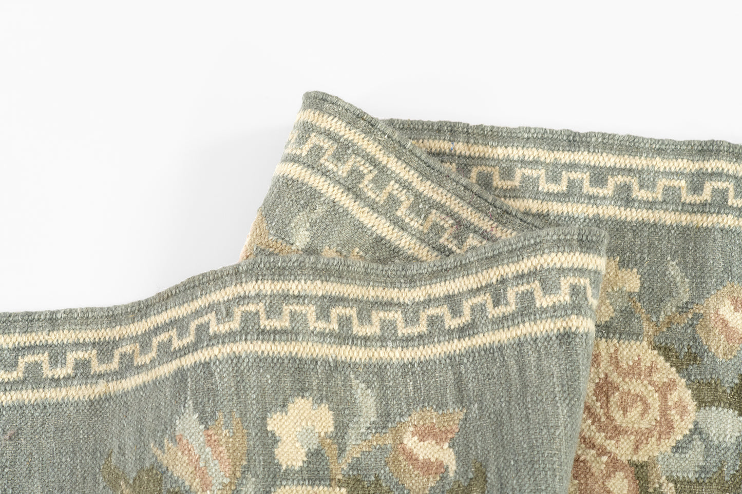 Blue-gray woven rug with cream floral pattern and Greek-key border, folded at the corner.