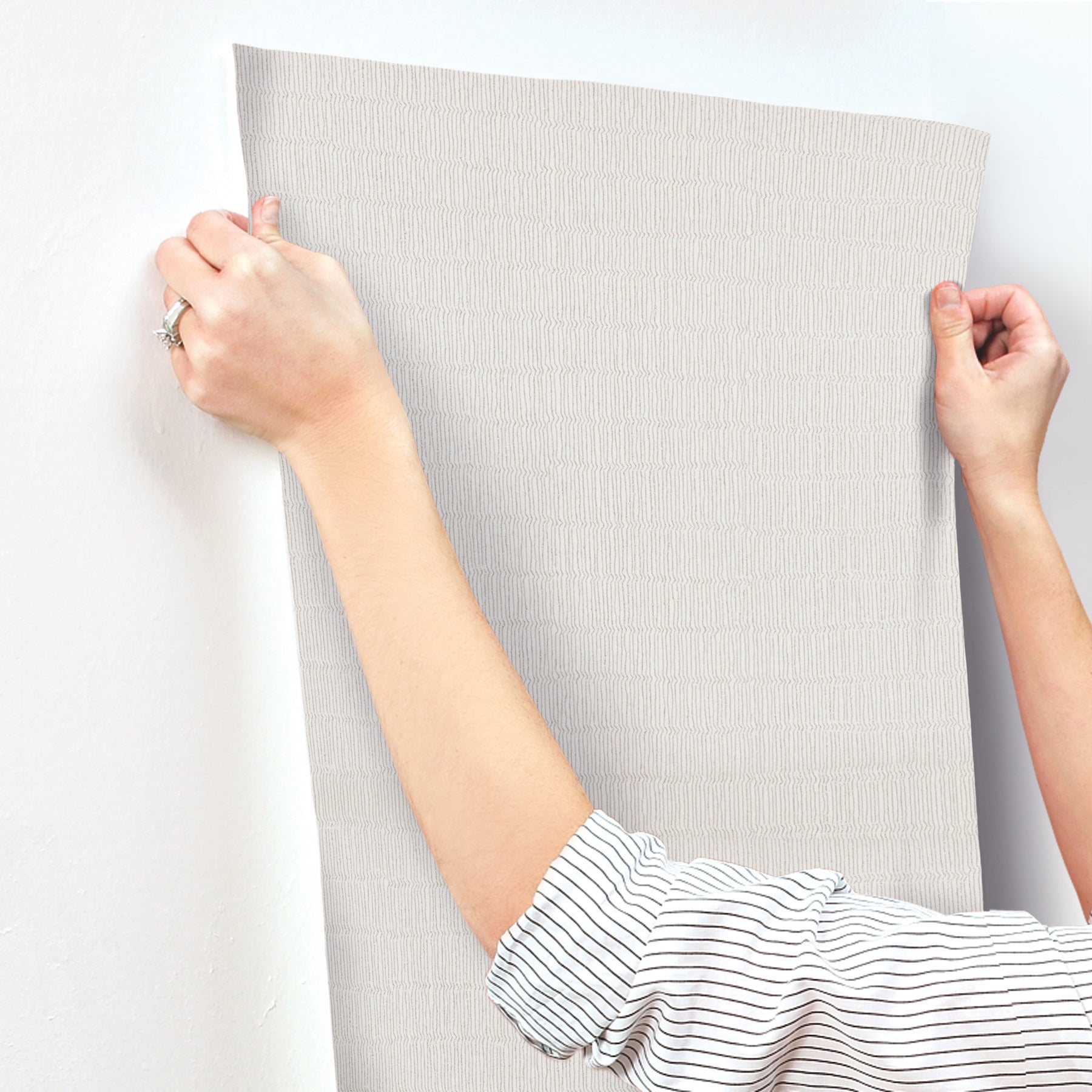 Light gray textured wallpaper sample, fabric-like with a subtle pattern, held up by two hands.