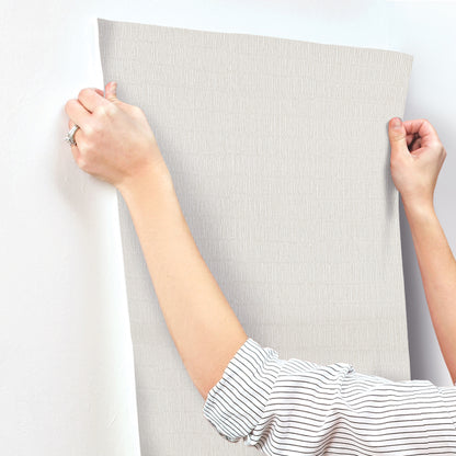 Light gray textured wallpaper sample, fabric-like with a subtle pattern, held up by two hands.