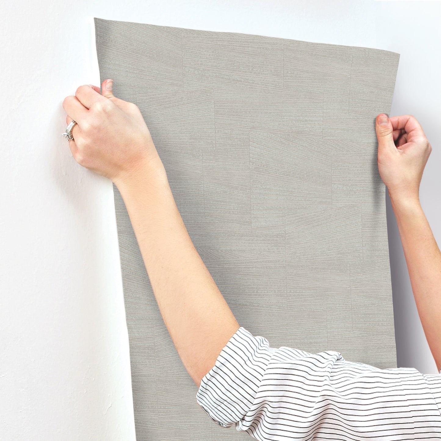 Hands hold a stone grey textured wallpaper panel with a geometric pattern against a wall; striped shirt sleeves visible.