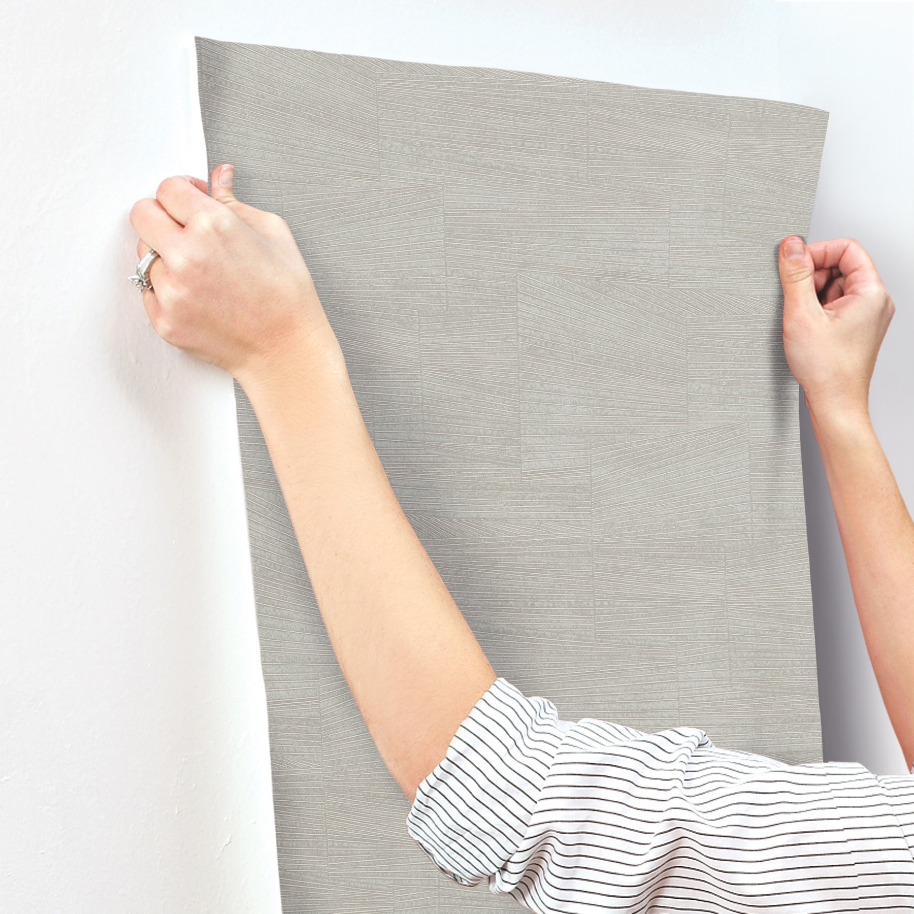 Hands hold a stone grey textured wallpaper panel with a geometric pattern against a wall; striped shirt sleeves visible.
