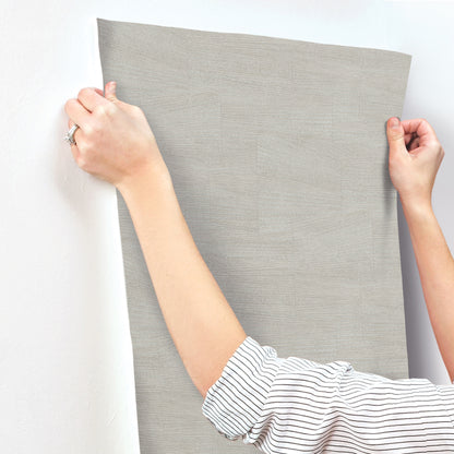 Hands hold a stone grey textured wallpaper panel with a geometric pattern against a wall; striped shirt sleeves visible.