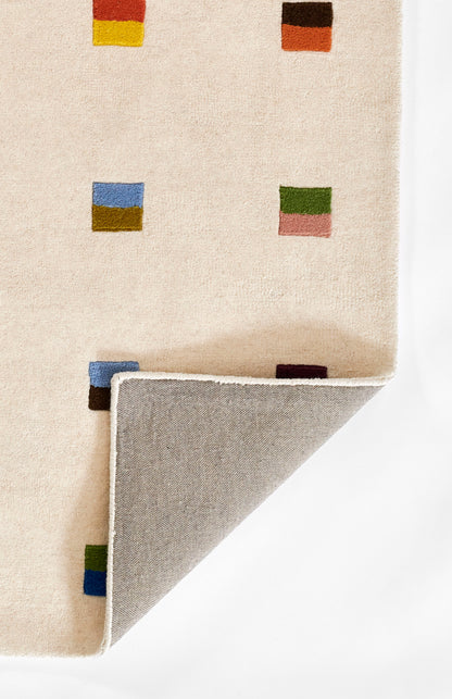 Beige rug with colorful square motifs; folded corner reveals gray underside and white binding.