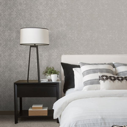 Bedroom wall with silver-grey mosaic tile wallpaper; black tripod bedside lamp, dark nightstand, white bedding, striped cushions.