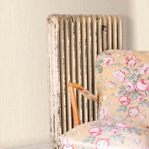 Cream plain-texture wallpaper backdrop with a distressed white vintage radiator behind a beige floral armchair with pink rose pattern and wooden arms.