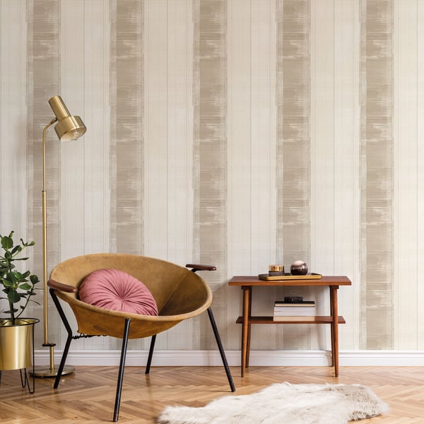 Beige striped wallpaper backdrop with a tan suede accent chair featuring a pink tufted cushion, a gold floor lamp, a wooden side table and greenery.