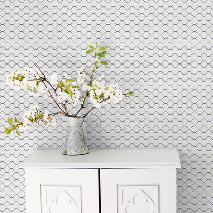 Blue geometric wallpaper backdrop behind a white two-door cabinet topped by a metal pitcher vase with white blossoms and branches.