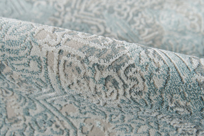 Close-up of blue-gray and beige damask rug with ornate floral pattern and woven texture.