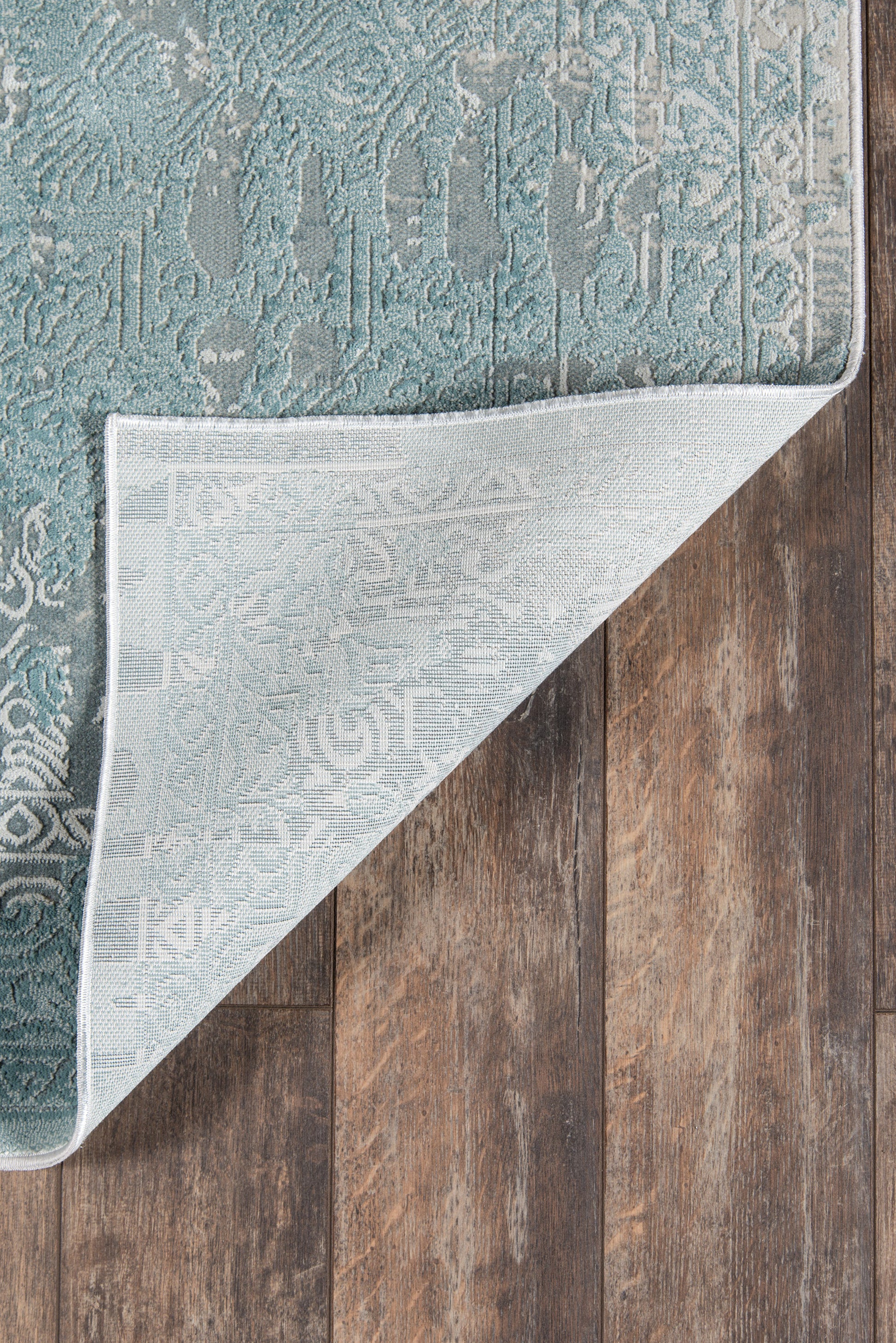 Teal patterned area rug with damask motif; corner flipped to show light underside on dark wood floor.