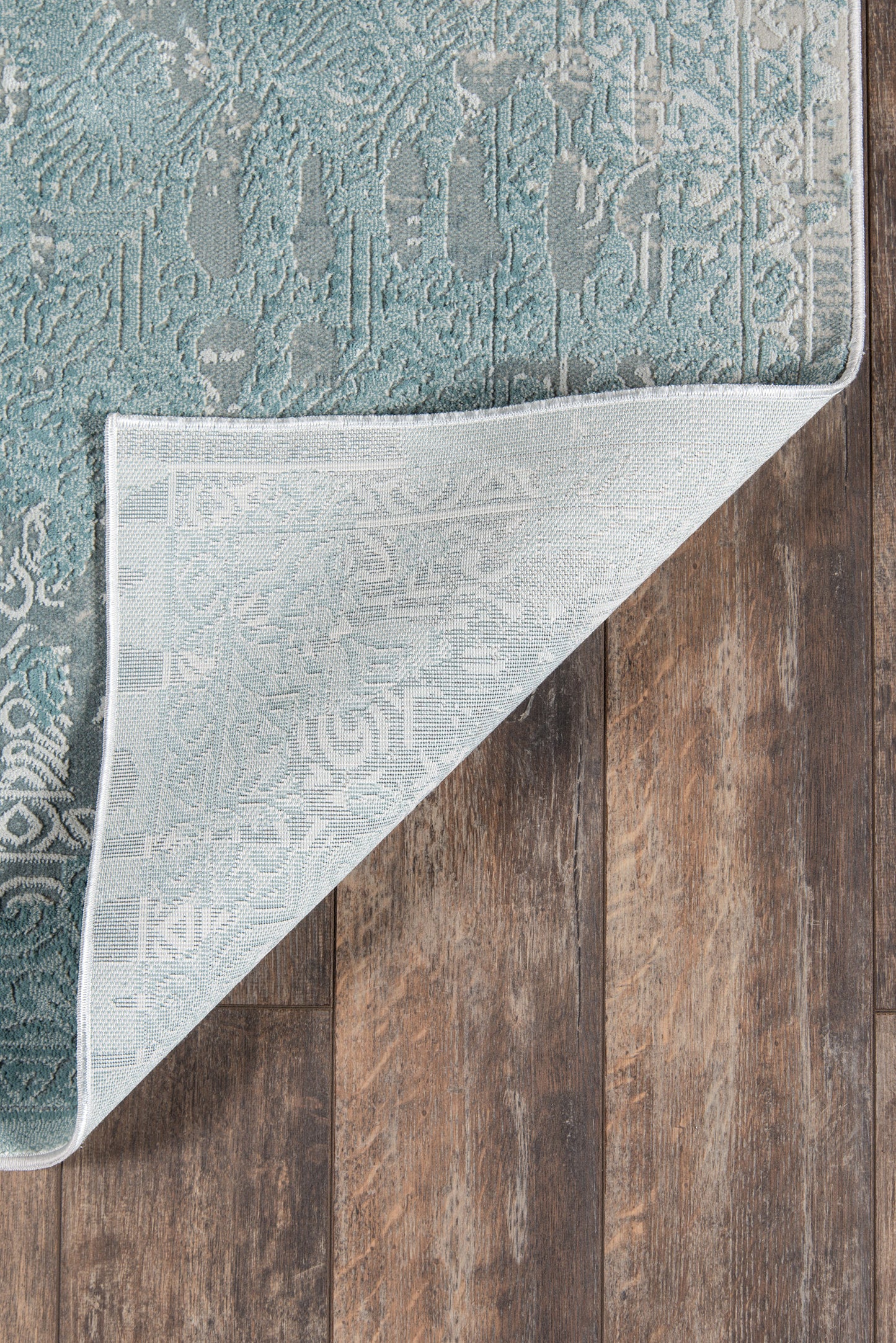 Teal damask patterned area rug with gray backing, folded corner on dark wood floor.