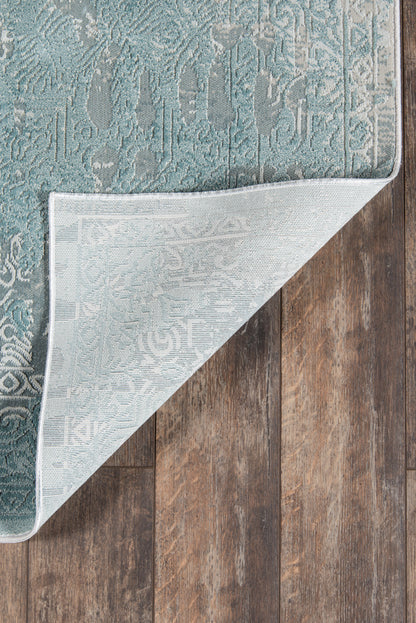 Teal damask patterned area rug with gray backing, folded corner on dark wood floor.