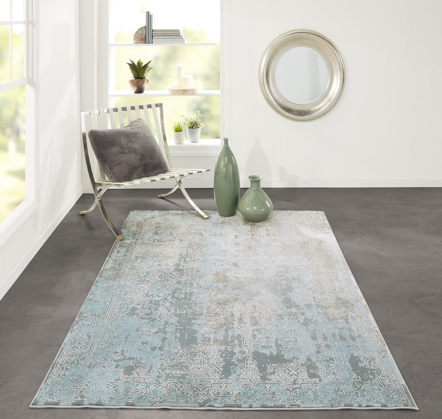 Blue-gray distressed rug with subtle border in a bright modern living room; green glass vases, white chair, round silver mirror.