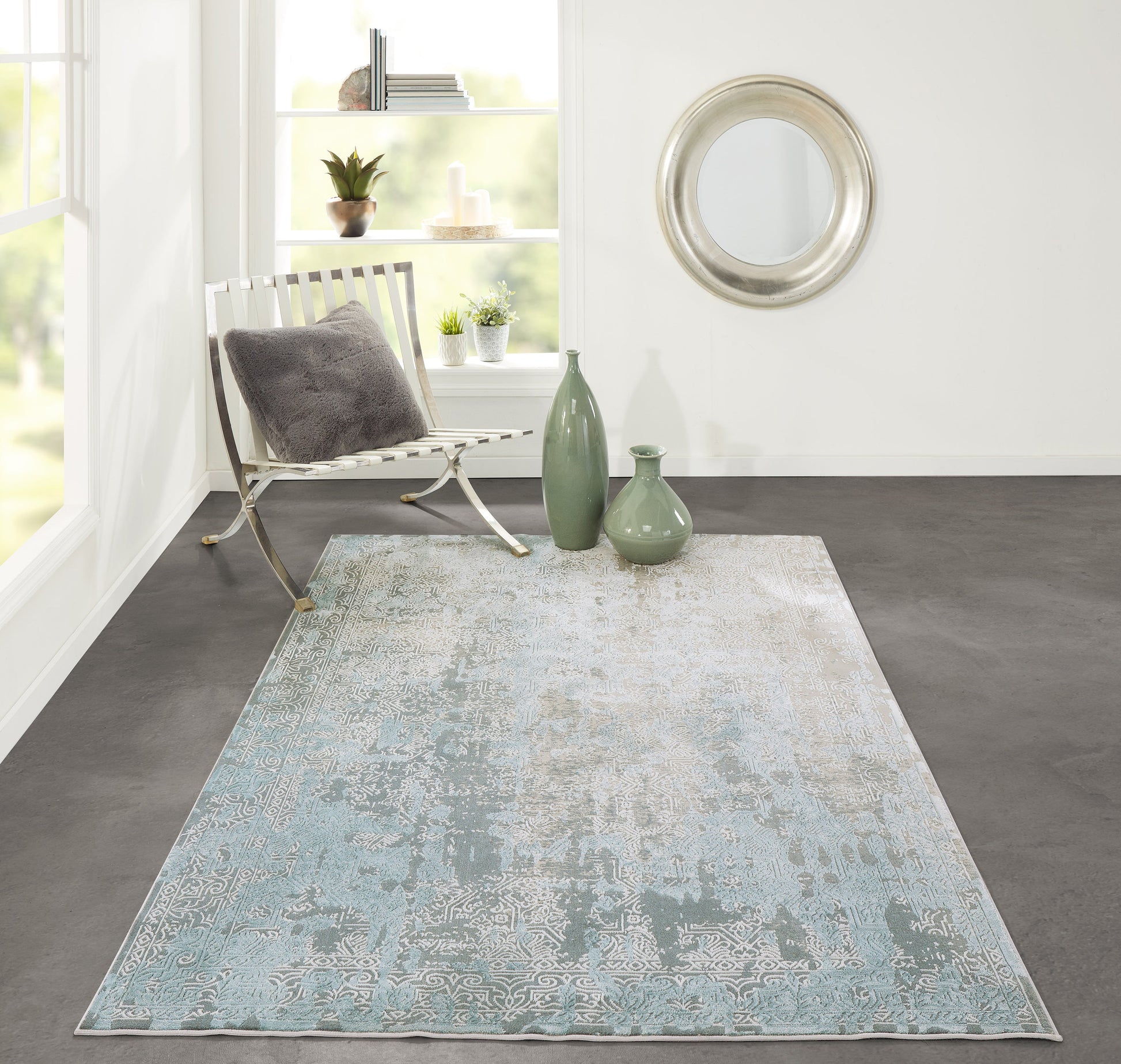 Modern living room with pale blue distressed rug, metal-frame chair, dark gray cushion & green vases, circular silver mirror.