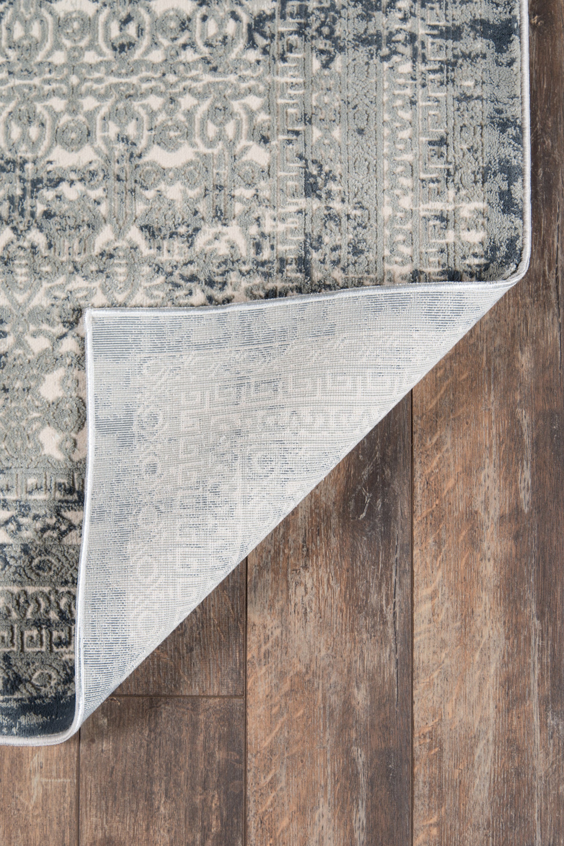 Blue-gray distressed patterned area rug with medallion motifs; folded corner reveals pale underside on dark wood floor.