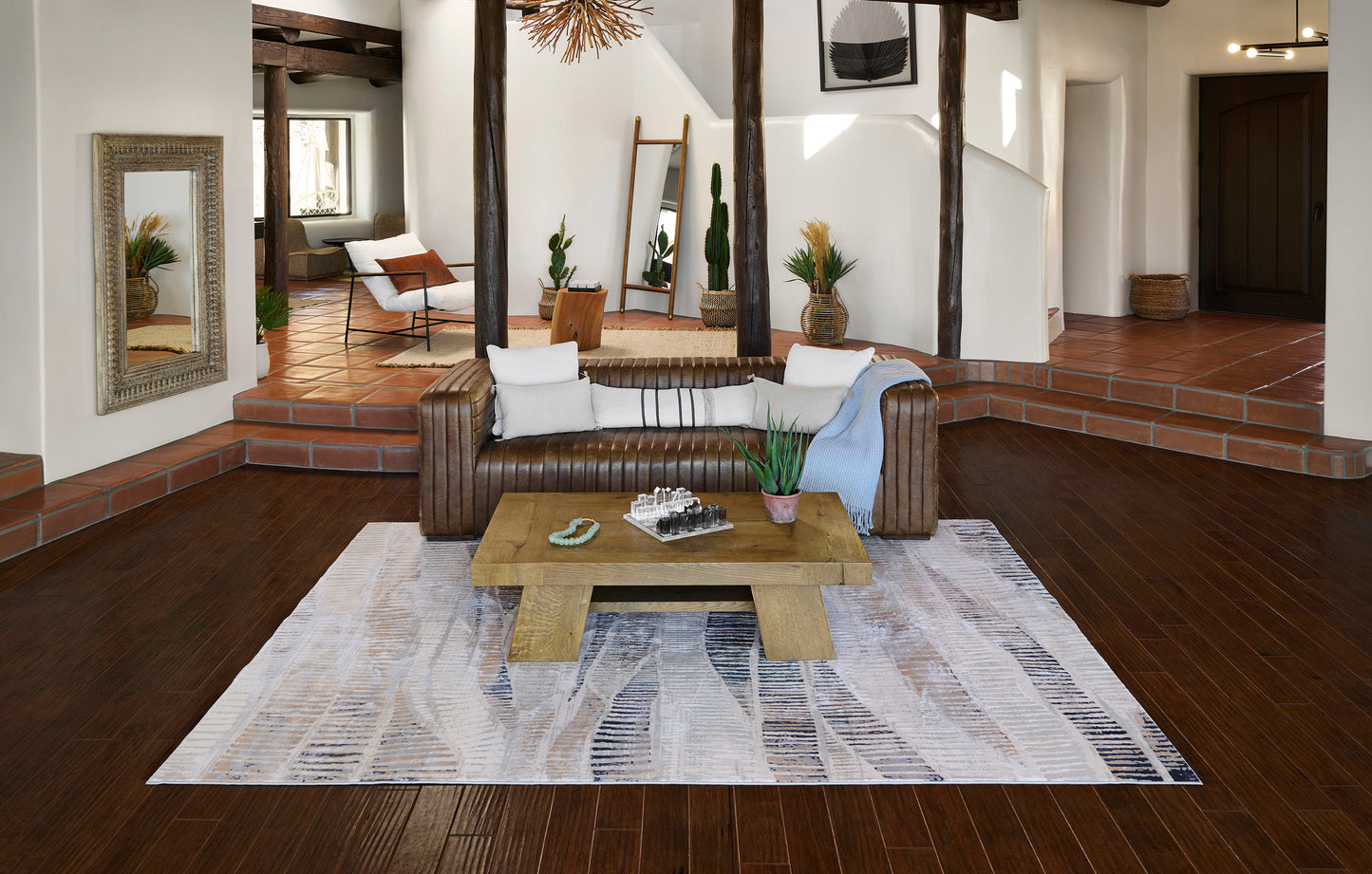 Rustic living room with dark wood floors, brown leather sofa, square wood coffee table, gray patterned rug, potted plants.