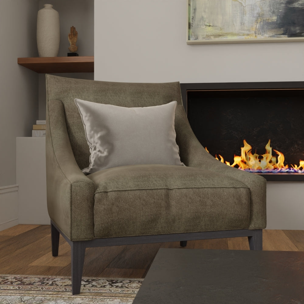 Olive Coco Solid fabric upholstery on an accent chair with dark legs and a light gray pillow in a living room by a lit fireplace.