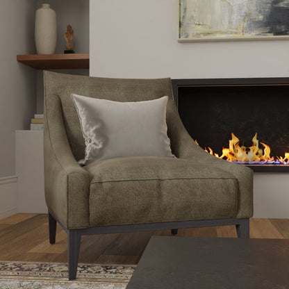 Olive Coco Solid fabric upholstery on an accent chair with dark legs and a light gray pillow in a living room by a lit fireplace.