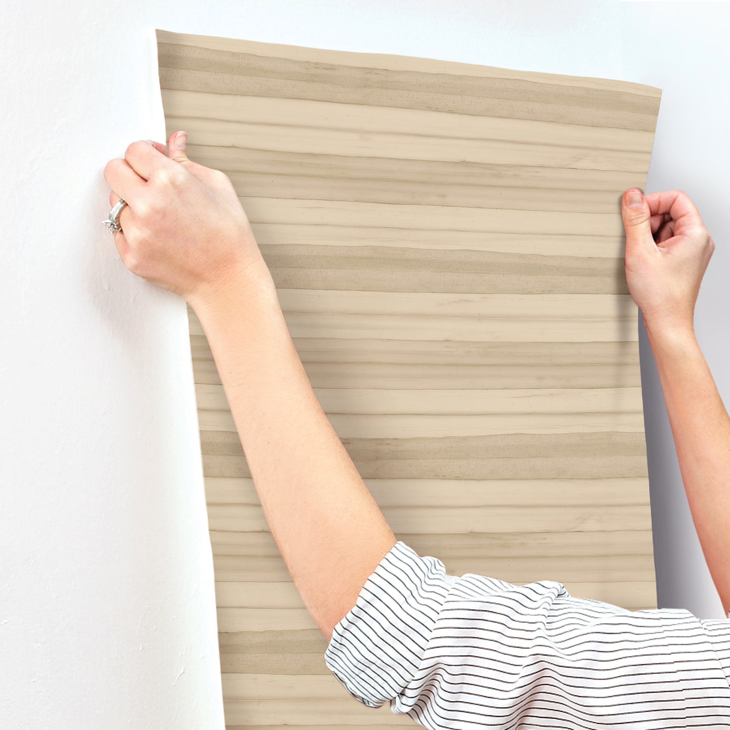 Two hands hold a pale striped wallpaper swatch against a white wall; casual striped shirt sleeve visible.
