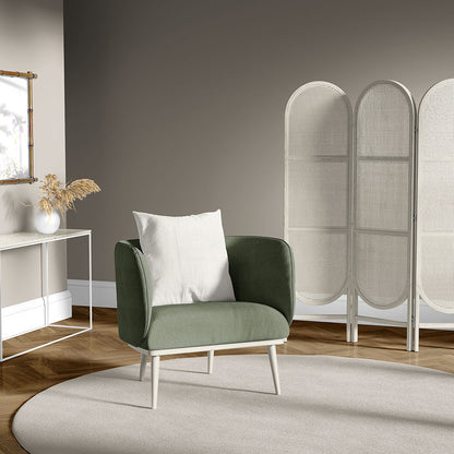 Olive green fabric armchair with light wood legs and white pillow, in a neutral living room with a woven room divider.