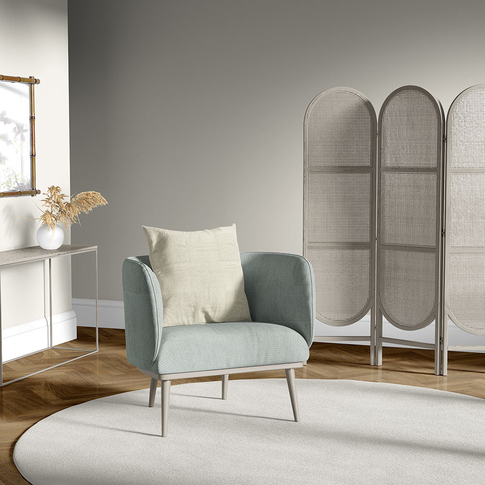 Soft blue-green upholstered armchair with light wooden legs and beige cushion, in minimalist living room with wicker screen.