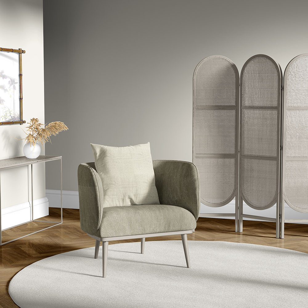 Olive-green fabric armchair with light cushion, wooden legs, beside a round rug and a wicker arched room divider.