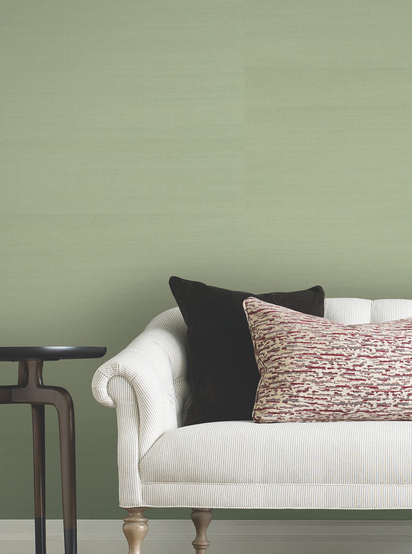 Soft sage grasscloth wallpaper on the wall behind a white textured sofa with rolled arms, black square cushion, and a red-beige patterned lumbar pillow; dark wood side table to the left.