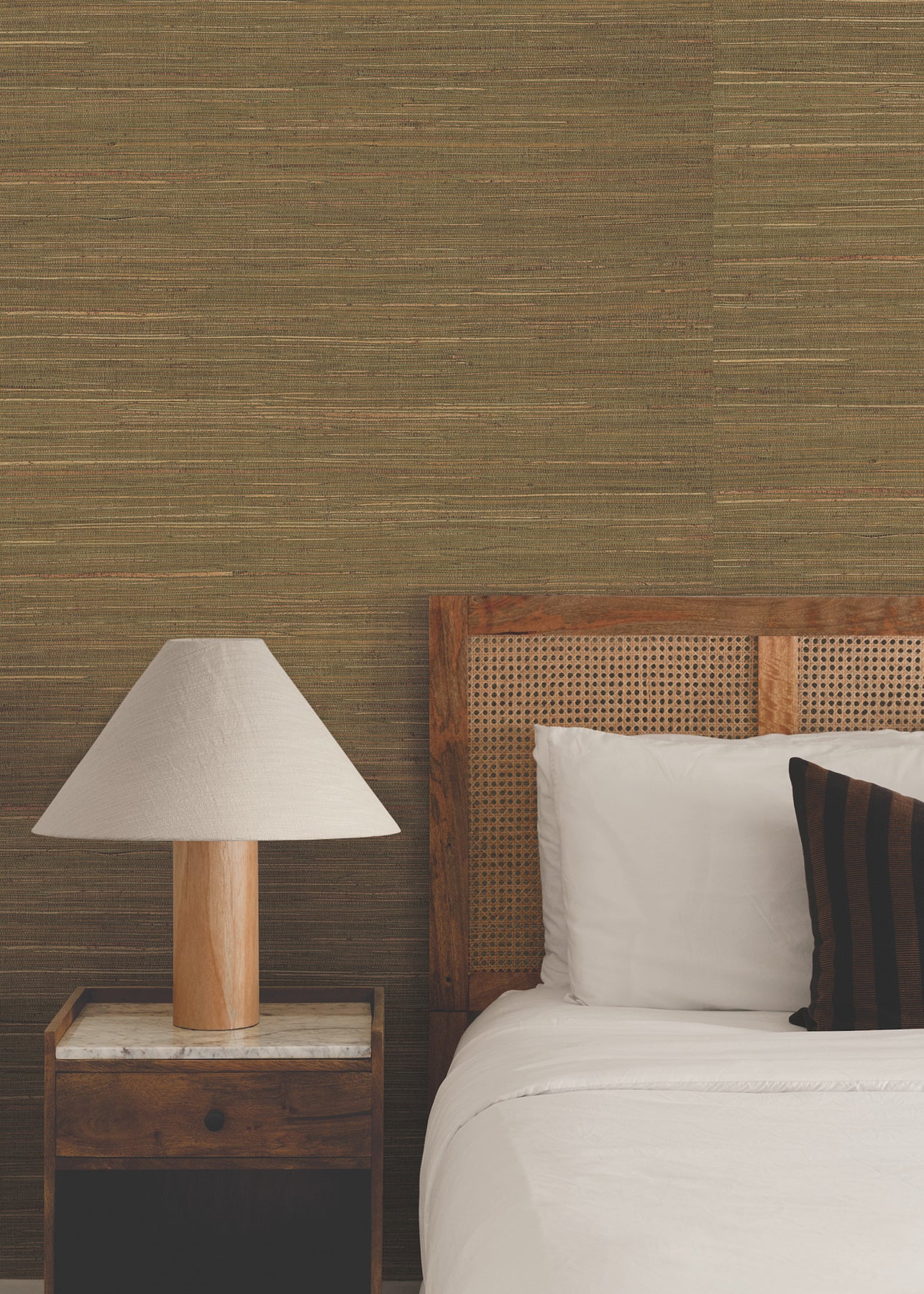 Bedroom wall covered in warm brown grasscloth wallpaper, with a cane headboard, white linens, dark striped pillow, wooden nightstand with marble top, and a lamp.