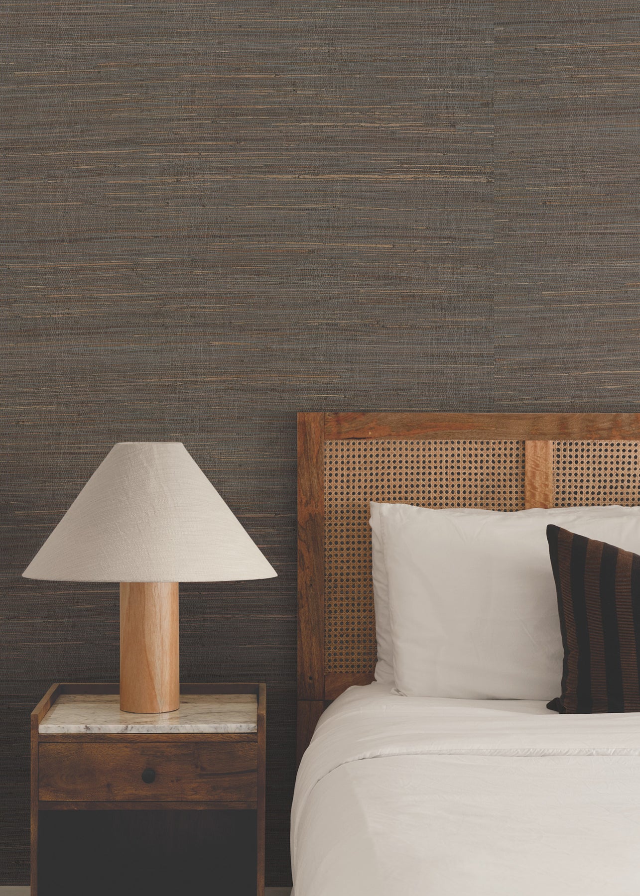 Cozy bedroom with caned wooden headboard, white bedding, dark striped pillow, marble-topped nightstand and wooden lamp.